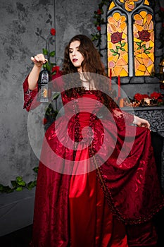 A beautiful girl in a magnificent red dress of the Rococo era stands against a fireplace, a window and flowers with a lamp with ca