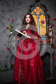 A beautiful girl in a magnificent red dress of the Rococo era stands with a book in her hands against the background of a fireplac