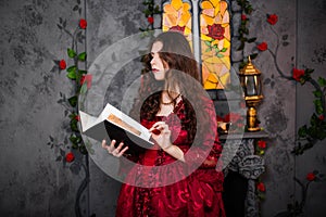 A beautiful girl in a magnificent red dress of the Rococo era stands with a book in her hands against the background of a fireplac