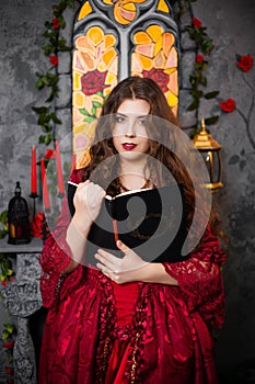 A beautiful girl in a magnificent red dress of the Rococo era stands with a book in her hands against the background of a fireplac