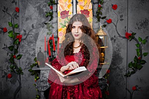 A beautiful girl in a magnificent red dress of the Rococo era stands with a book in her hands against the background of a fireplac