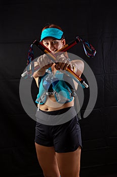 Beautiful girl marathon runner in blue jogging equipment and trekking poles in the studio on a black background