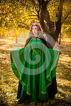 A beautiful girl in a medieval green dress with golden braid. Queen in a cloak and a blio dress in the autumn forest