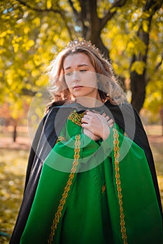 A beautiful girl in a medieval green dress with golden braid. Queen in a cloak and a blio dress in the autumn forest
