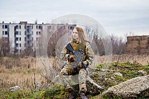 A beautiful girl in military uniform with an airsoft gun sitting on a mossy stone heap with ruined buildings in the background