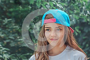 Beautiful girl model in a baseball cap with flowing hair on the background of green shrubs or trees. Portrait of a happy