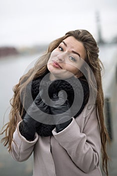 Beautiful girl in a natural make-up cute smiling at the camera. Girl in a black coat, a scarf and a red dress against a gray sky