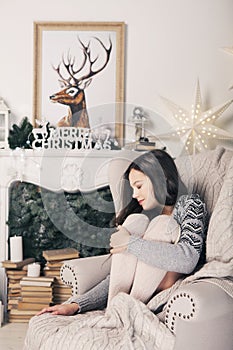 Beautiful girl near fireplace with Christmas tree, candles sits in comfortable armchair. Scandinavian cozy home decor