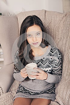 Beautiful girl near fireplace with Christmas tree, candles sits in comfortable armchair. Scandinavian cozy home decor