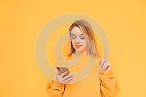 Beautiful girl in orange clothes uses a smartphone on a yellow background and shows her finger up to a blank space. Girl is