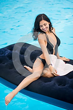 Beautiful girl with perfect figure in a black bikini and hat sitting on a mattress in the swimming pool