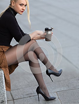 Beautiful girl with perfect legs in pantyhose sitting outdoor on the city street with coffee thermos mug