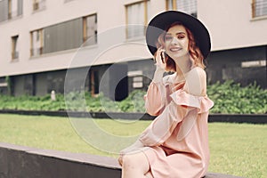 Beautiful girl in a pink dress sitting near high-rise buildings . Girl speaks by phone. Woman with pink hair in a black hat