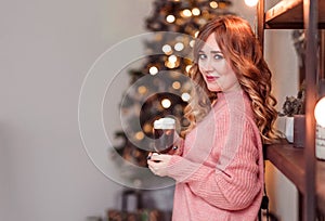 Beautiful girl in a pink sweater with a cup of coffee and marshmallows in hands on a background of New Year  lights