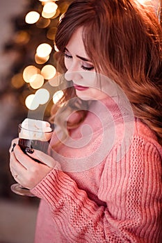 Beautiful girl in a pink sweater with a cup of coffee and marshmallows in hands on a background of New Year  lights