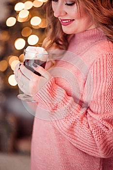 Beautiful girl in a pink sweater with a cup of coffee and marshmallows in hands on a background of New Year  lights