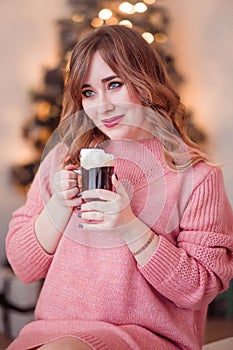 Beautiful girl in a pink sweater with a cup of coffee and marshmallows in hands on a background of New Year  lights