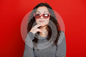 Beautiful girl portrait posing and flirting on red background, long curly hair, sunglasses in heart shape, glamour concept