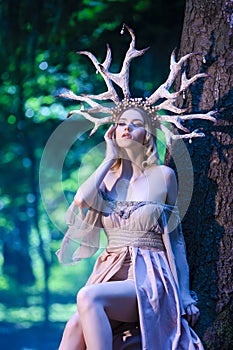 Beautiful Girl Posing With Artistic Deer Horns  In Summer Forest Near High Tree. Wearing Light Dress for Forest Nymph Concept. Art