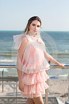 A beautiful girl posing on a transparent balcony on a blue sea background. A lady in a pink dress with ruches near the sea.