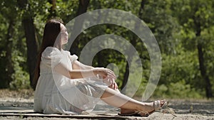 A beautiful girl in positive emotions sits sideways on the sand in a summer forest and meditates. A lady in a white