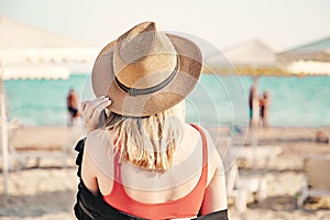 Beautiful girl in a red bikini and straw hat on the beach. Back view. White sand, blue water and beach umbrellas on the