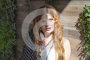 A beautiful girl with red curly hair stands at the wooden wall around the plan in the sunlight.