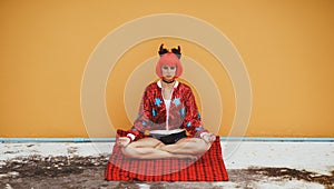 Beautiful girl in red devil costume sitting in yoga pose in anticipation of Halloween. Wears a red wig and horns. The