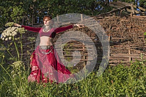 Beautiful girl in red dress dances incendiary dance on summer meadow against of hedge.