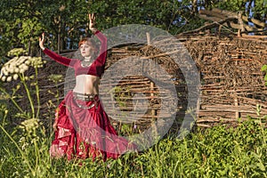 Beautiful girl in red dress dances incendiary dance on summer meadow against of hedge.