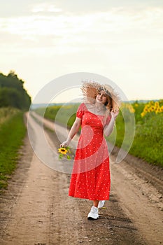 A beautiful girl in a red dress and hat is walking along a rural dirt road.