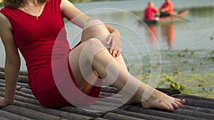 Beautiful girl in red dress is sitting on wooden pier. Woman is embracing her knees near the lake. Rustic and natural photo