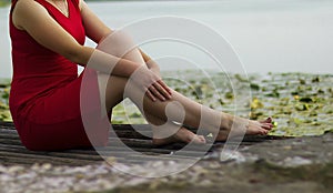 Beautiful girl in red dress is sitting on wooden pier. Woman is embracing her knees near the lake. Rustic and natural photo