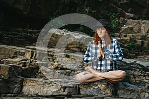 Beautiful girl with red hair in a hat and shirt meditating on rocks in a lotus position. Space for your text message or