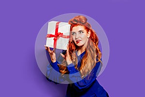 A beautiful girl with red hair and red lipstick in a blue dress in the style of pin-up on a purple background holds a gift and