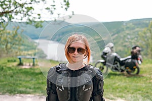 Beautiful girl with red hair and sunglasses posing on a motorcycle background. Dressed in a turtle jacket, body armor. Nature and