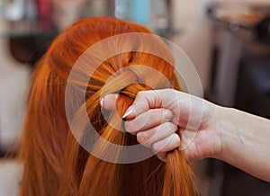 Beautiful girl with red long hair, hairdresser weaves a French braid, in a beauty salon.