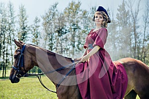 Beautiful girl in a red long red dress and in a black hat with a cocked hat riding a brown horse.