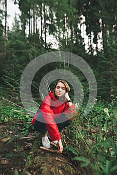 Beautiful girl in a red raincoat sits on a log over a mountain river, looks into the camera with a serious face. Portrait of a