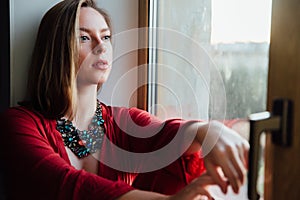 Beautiful girl in a red robe is sitting on the windowsill at home.
