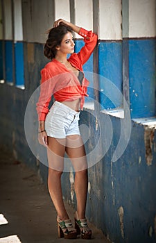 Beautiful girl with red shirt and white shorts posing in old hall with columns blue painted. Attractive long hair brunette