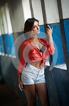 Beautiful girl with red shirt and white shorts posing in old hall with columns blue painted. Attractive long hair brunette