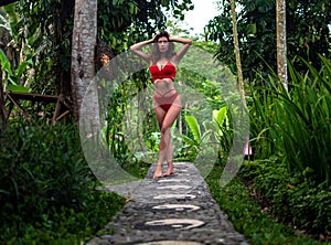 Beautiful girl in red swimsuit posing in tropical location with green trees. Young sports model in bikini with perfect