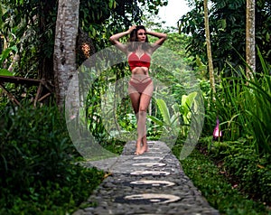 Beautiful girl in red swimsuit posing in tropical location with green trees. Young sports model in bikini with perfect