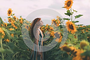 Beautiful girl with red wavy hair and freckles in stripped colourful dress enjoying nature on the field of sunflowers.