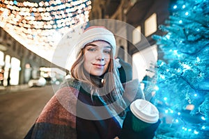 Beautiful girl in a Santa hat stands near a Christmas tree on a street background decorated with Christmas lights, holds a cup of
