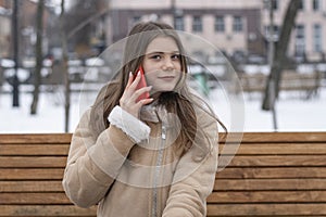 Beautiful girl in sheepskin coat sits on bench in winter park and talks on smartphone. Serious young woman with gadget