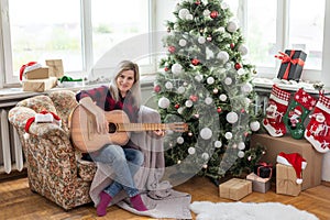 beautiful girl sits near the Christmas tree decorated with balls and light and playing the guitar and singing. Young