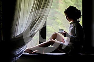 A beautiful girl sits on the windowsill near a large window and drinks her morning coffee