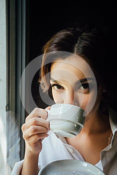 A beautiful girl sits on the windowsill near a large window and drinks her morning coffee